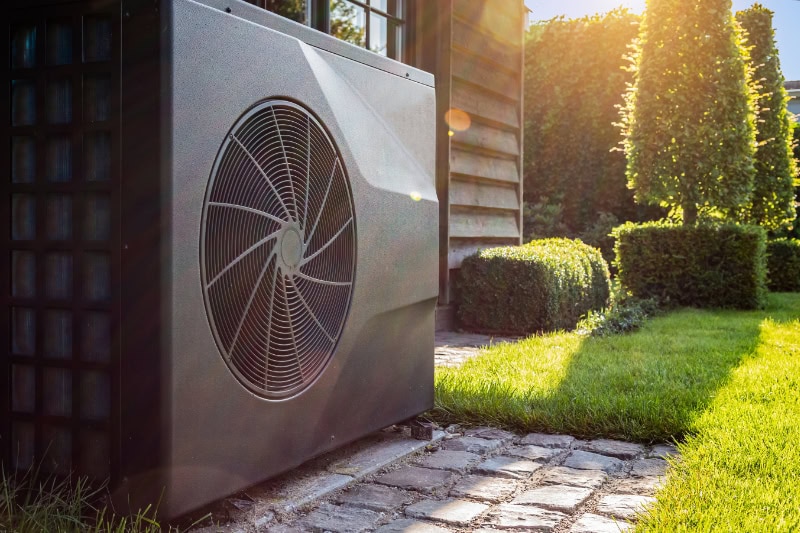 Close-up of black full inverter heat pump outside in the garden, near wooden pool house on a sunny day.